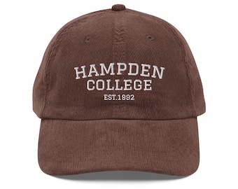 Puede incluir: Gorra de béisbol de pana marrón con el texto blanco "HAMPDEN COLLEGE" y "EST.1992". La gorra tiene una visera curva y una parte superior redondeada. Un sombrero de estilo clásico.