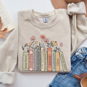 Puede incluir: Una sudadera beige con un gráfico de una pila de libros con acentos florales. Los títulos de los libros incluyen "Jane Eyre", "Fourth Wing", "Mansfield Park", "The Book Thief", "Pride and Prejudice", "The Kite Runner", "Beloved", "Frankenstein", "Circe" y "Little Women".