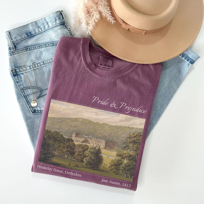 Puede incluir: Una camiseta malva con las palabras "Pride & Prejudice" y una imagen de paisaje de Pemberley House, Derbyshire. La camiseta se combina con vaqueros azul claro y un sombrero beige con una cinta marr&oacute;n.