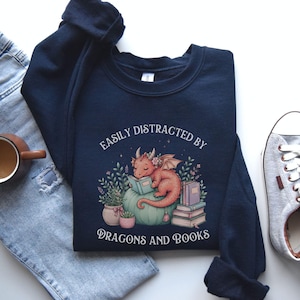 May include: A navy blue crewneck sweatshirt featuring a graphic of a small dragon reading a book while perched on a large pumpkin. The text reads EASILY DISTRACTED BY DRAGONS AND BOOKS, surrounded by potted plants and a stack of books in soft colors.