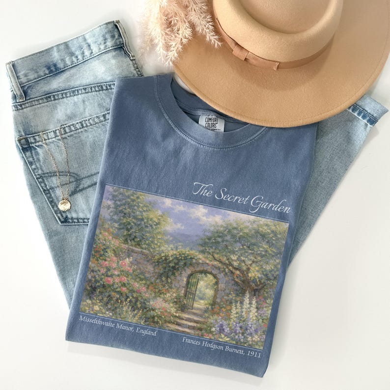 May include: A light blue t-shirt with the text "The Secret Garden" and a landscape painting. The image also includes a pair of light wash jeans, a tan hat, and a gold necklace.