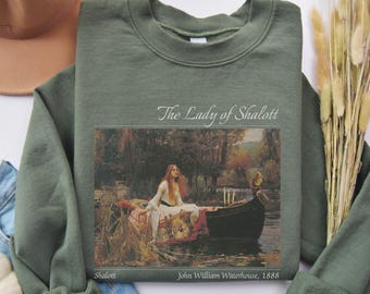 Puede incluir: Sudadera verde oliva con una impresión de la pintura "The Lady of Shalott" de John William Waterhouse. La obra de arte representa a una mujer en un bote en un río. El texto incluye "The Lady of Shalott", "Shalott" y "John William Waterhouse, 1888."