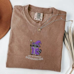 Op de afbeelding: Een bruin Comfort Colors T-shirt met een geborduurde wasbeer met een paarse tovenaarsmantel en -hoed, met een staf. De tekst "OFF TO COMMIT SHENANIGANS" staat onder de wasbeer. Een zilveren ketting met een hanger ligt op het shirt.