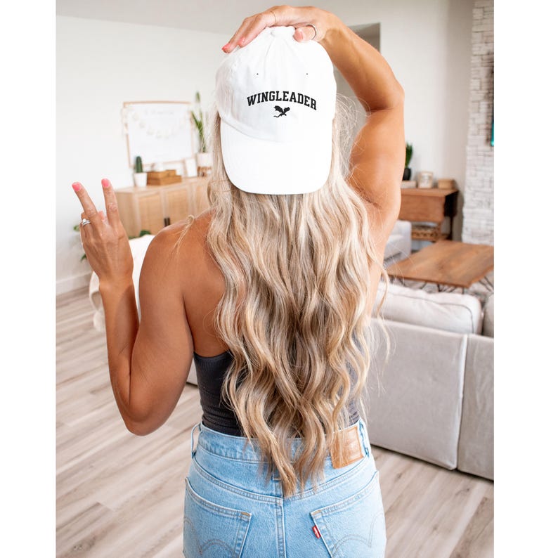 May include: White baseball cap with black embroidery that reads "WINGLEADER" with a small graphic of a wing. The cap is worn by a person with long blonde hair.