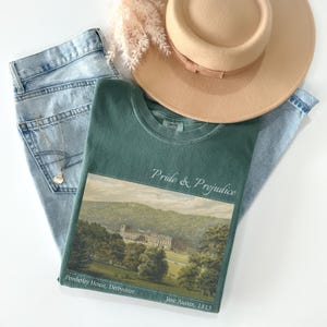 Puede incluir: Una camiseta verde con las palabras "Pride & Prejudice" y una imagen de paisaje de Pemberley House, Derbyshire. La camiseta se combina con vaqueros azul claro, un sombrero beige y un collar dorado.