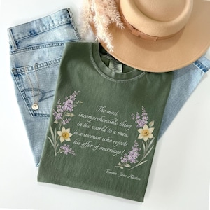Può includere: Una t-shirt verde oliva con una citazione stampata di Jane Austen, circondata da illustrazioni di fiori di lillà viola e narcisi gialli. Il testo recita, The most incomprehensible thing in the world to a man, is a woman who rejects his offer of marriage! Emma - Jane Austen.