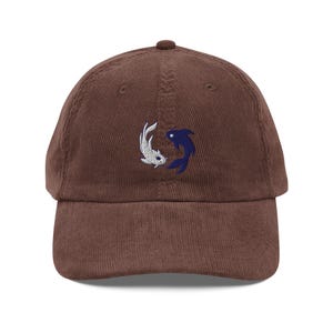 May include: A brown corduroy baseball cap featuring an embroidered design of two koi fish, one white and one blue, in a yin-yang arrangement. The cap has a curved brim and an adjustable strap at the back. The hat is a casual accessory.