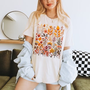 May include: A white t-shirt with a colorful floral print featuring various shades of orange, yellow, red, and green. The print is a square design with a variety of flowers, leaves, and stems.