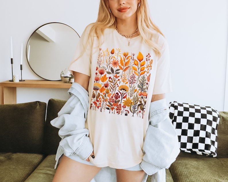May include: A white t-shirt with a colorful floral print featuring various shades of orange, yellow, red, and green. The print is a square design with a variety of flowers, leaves, and stems.