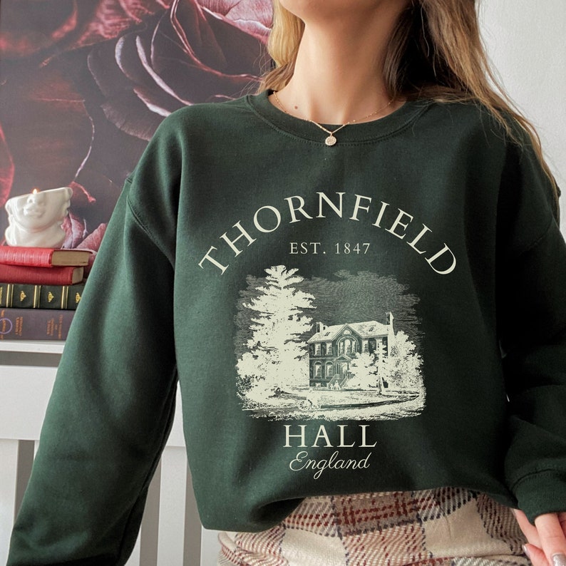 May include: A dark green sweatshirt with a white graphic print of a house and trees. The text "Thornfield Hall, Est. 1847, England" is printed on the sweatshirt.