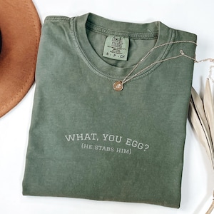 Könnte beinhalten: Ein olivgrünes T-Shirt mit dem Text "WHAT, YOU EGG? (HE STABS HIM)". Das Shirt hat einen Rundhalsausschnitt und kurze Ärmel. Eine goldene Halskette mit Anhänger liegt über dem Shirt.