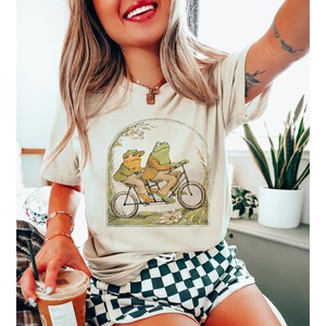 K&ouml;nnte beinhalten: Ein wei&szlig;es T-Shirt mit einem Aufdruck von zwei Fr&ouml;schen, die ein Tandem fahren. Die Fr&ouml;sche tragen H&uuml;te und das Fahrrad ist von gr&uuml;nem Laub umgeben.