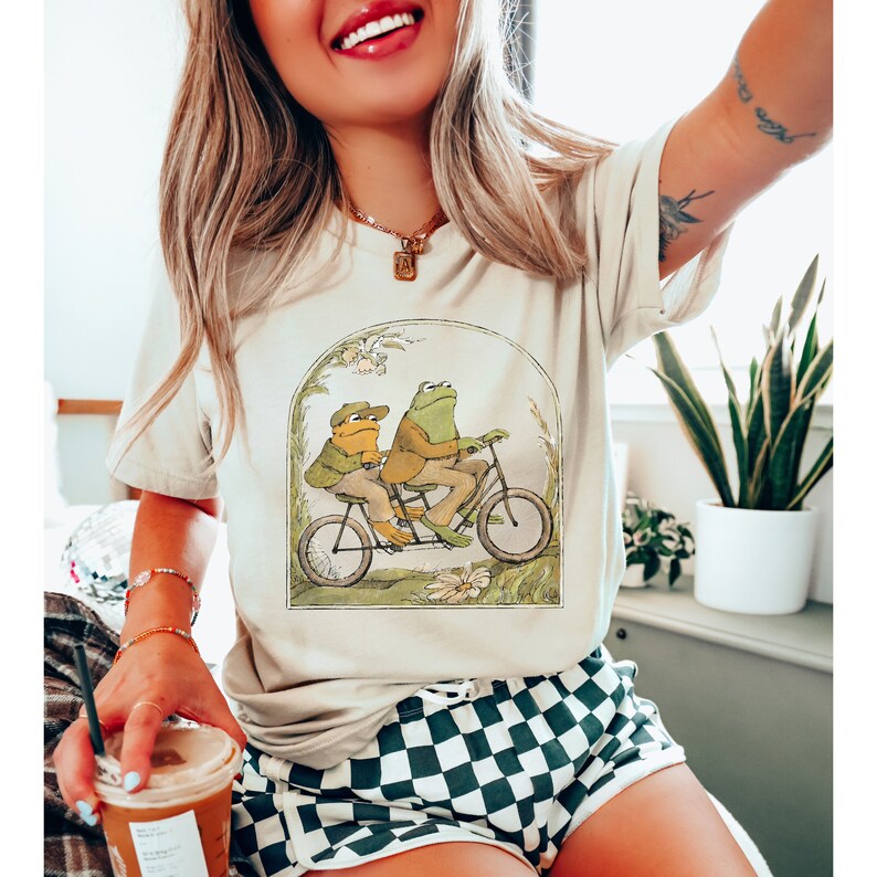 K&ouml;nnte beinhalten: Ein wei&szlig;es T-Shirt mit einem Aufdruck von zwei Fr&ouml;schen, die ein Tandem fahren. Die Fr&ouml;sche tragen H&uuml;te und das Fahrrad ist von gr&uuml;nem Laub umgeben.