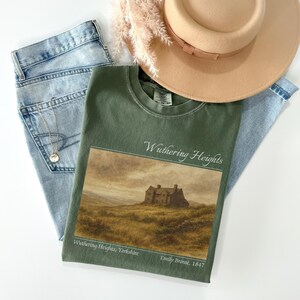 Op de afbeelding: Een olijfgroen t-shirt met een landschapsafbeelding van een huis op een heuvel en de tekst "Wuthering Heights". Het shirt wordt gecombineerd met een lichtblauwe jeans, een gouden ketting en een beige hoed.