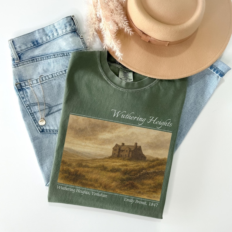 Op de afbeelding: Een olijfgroen t-shirt met een landschapsafbeelding van een huis op een heuvel en de tekst "Wuthering Heights". Het shirt wordt gecombineerd met een lichtblauwe jeans, een gouden ketting en een beige hoed.
