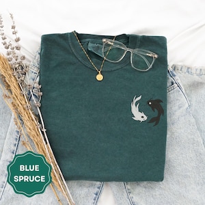 May include: A blue spruce t-shirt with a Yin Yang koi fish design. The shirt has a crew neck and short sleeves. A gold necklace and clear glasses rest on the shirt. The shirt is displayed on a pair of blue jeans.