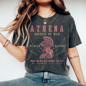 May include: A dark grey t-shirt with a vintage-style graphic of Athena, the Greek goddess of war. The graphic features a pink illustration of Athena wearing a helmet and wings, with the text "ATHENA, GODDESS OF WAR, WISDOM, WARTIME, WHEN THE OBSTACLE BECOMES THE WAY, MT. OLYMPUS 9.26.1217BC".