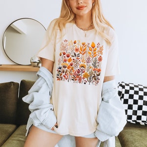 Może przedstawiać: Biały t-shirt z nadrukiem kwiatowym przedstawiającym r&oacute;żne kwiaty i liście w odcieniach pomarańczowego, ż&oacute;łtego, czerwonego i zielonego. Koszulka ma luźny kr&oacute;j i okrągły dekolt.