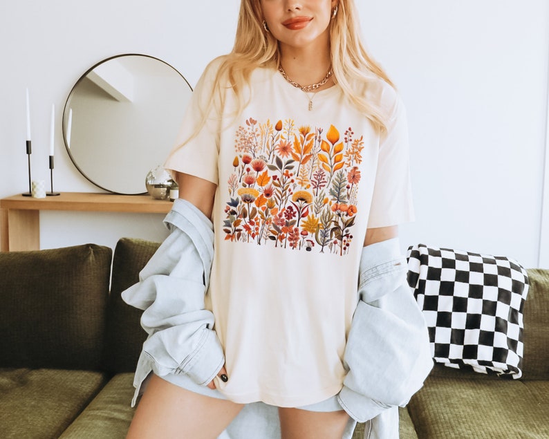 Może przedstawiać: Biały t-shirt z nadrukiem kwiatowym przedstawiającym r&oacute;żne kwiaty i liście w odcieniach pomarańczowego, ż&oacute;łtego, czerwonego i zielonego. Koszulka ma luźny kr&oacute;j i okrągły dekolt.