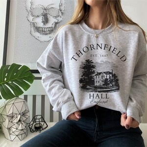 May include: Gray sweatshirt with a black graphic print of a house and the text "Thornfield Hall, Est. 1847, England".