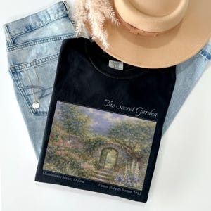 May include: Black t-shirt featuring a landscape painting with the text "The Secret Garden." The image includes a light brown hat, light blue jeans, and a gold necklace. The painting depicts a garden scene with an arched doorway.