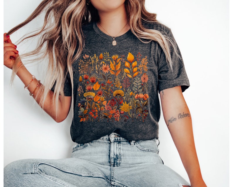 Może przedstawiać: Szary t-shirt z nadrukiem kwiatowym z ż&oacute;łtymi, pomarańczowymi i czerwonymi kwiatami. Kwiaty są ułożone w pionowy wz&oacute;r i otoczone zielonymi liśćmi.