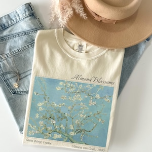 Puede incluir: Camiseta color crema con la pintura "Almendros en flor" de Van Gogh. La obra representa flores blancas sobre un cielo azul. La camiseta se combina con vaqueros de lavado claro y un sombrero beige.