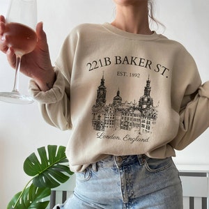 Può includere: Una felpa beige con una stampa grafica nera di un edificio con il testo "221B Baker St. Est. 1892 Londra, Inghilterra" sul davanti.