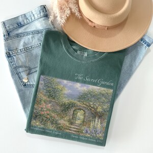 May include: An overhead shot features a green t-shirt with "The Secret Garden" text and a scenic print, paired with light-wash jeans, a tan hat, and a gold necklace. The shirt's print depicts a garden scene with a stone archway.