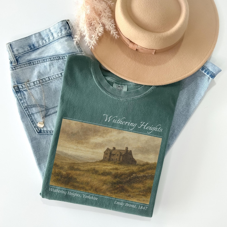 Op de afbeelding: Een casual mode flatlay met een teal groen t-shirt met een "Wuthering Heights" afbeelding, lichtblauwe jeans, een bruine fedora hoed en een gouden ketting. Het t-shirt heeft de tekst "Wuthering Heights, Yorkshire, Emily Bront&euml;, 1847."