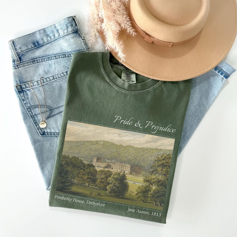 Puede incluir: Una camiseta verde oliva con el texto "Pride & Prejudice" y una imagen de paisaje de Pemberley House, Derbyshire. La camiseta se combina con unos vaqueros de lavado claro, un collar dorado y un sombrero beige con una banda marr&oacute;n.