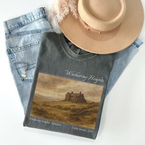 Op de afbeelding: Een grijs t-shirt met de tekst "Wuthering Heights" en een afbeelding van een huis op een heuvel. Het shirt wordt gecombineerd met lichtblauwe jeans en een beige hoed. Een zilveren ketting is zichtbaar.