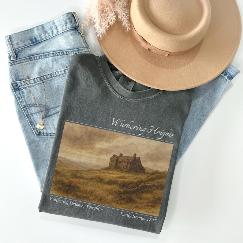 Op de afbeelding: Een grijs t-shirt met de tekst "Wuthering Heights" en een afbeelding van een huis op een heuvel. Het shirt wordt gecombineerd met lichtblauwe jeans en een beige hoed. Een zilveren ketting is zichtbaar.