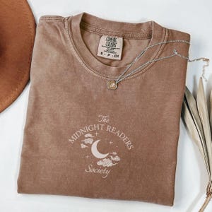 Peut inclure: Un t-shirt Comfort Colors marron avec le texte "The Midnight Readers Society" en blanc. Un collier argenté avec un pendentif est drapé sur le t-shirt. Un chapeau marron et des plantes séchées sont en arrière-plan.