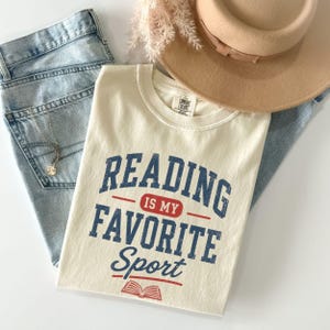 May include: A cream-colored short-sleeve t-shirt featuring the blue and red text "READING IS MY FAVORITE Sport" with a small book icon at the bottom. The shirt is layered over light blue denim jeans and sits beneath a tan wide-brimmed hat.