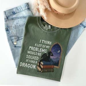 Könnte beinhalten: Ein olivgrünes T-Shirt mit einer Grafik eines schwarzen Drachen, der auf einem Stapel von drei Büchern sitzt. Der Text lautet, I THINK A LOT OF MY PROBLEMS WOULD BE SOLVED IF I HAD A DRAGON. Neben den Büchern ist ein Bogenfenster mit nächtlichem Himmel zu sehen.
