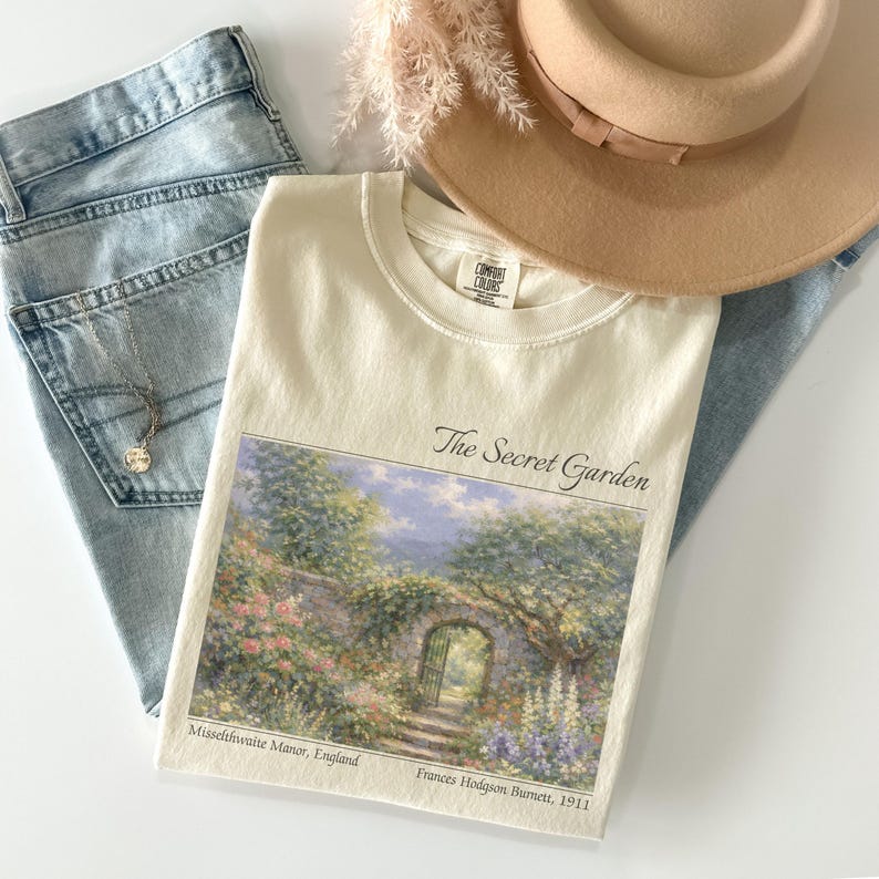 May include: Cream-colored t-shirt with "The Secret Garden" graphic, jeans, a necklace, and a tan hat. The graphic depicts a garden scene with a stone archway and flowers. The text "Misselthwaite Manor, England" and "Frances Hodgson Burnett, 1911" are also visible.