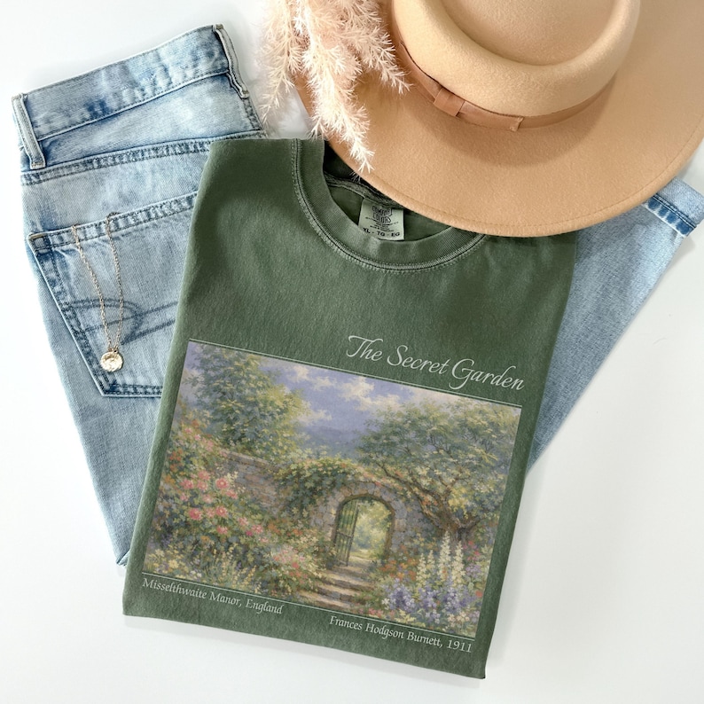 May include: An olive green t-shirt with a landscape print of a garden scene, the text "The Secret Garden", and light blue jeans. A tan hat and a gold necklace are also in the image.