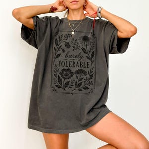 May include: A charcoal grey short-sleeve t-shirt featuring a black rectangular frame containing botanical illustrations of flowers and leaves, with the text "barely TOLERABLE" printed in the centre in a serif font.