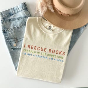 May include: A cream-colored t-shirt with the text I RESCUE BOOKS TRAPPED IN THE BOOKSTORE. I'M NOT A HOARDER, I'M A HERO. printed in red, green, and blue. The shirt is paired with light blue denim jeans, a tan wide-brimmed hat, and a gold necklace.