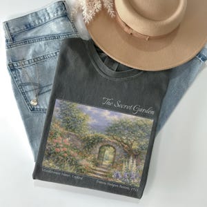 May include: A casual ensemble featuring a dark gray t-shirt with "The Secret Garden" artwork, light wash jeans, a tan hat, and a delicate necklace. The t-shirt displays a scenic garden illustration. The hat has a brown band.