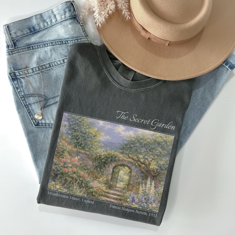 May include: A casual ensemble featuring a dark gray t-shirt with "The Secret Garden" artwork, light wash jeans, a tan hat, and a delicate necklace. The t-shirt displays a scenic garden illustration. The hat has a brown band.