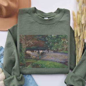 Op de afbeelding: Een olijfgroene sweater met ronde hals en een rechthoekige grafische print van een persoon die in een beek drijft, omringd door weelderig groen en bloemen, met de witte tekst "I'm literally fine" gecentreerd over de afbeelding.