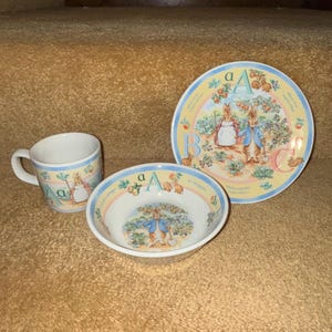 May include: A Peter Rabbit themed children's dish set with a blue and white bowl, a blue and white mug, and a blue and white plate. The plate features the letter A and the text "A is for apple." The bowl and mug feature the letter A and a rabbit holding a spade.