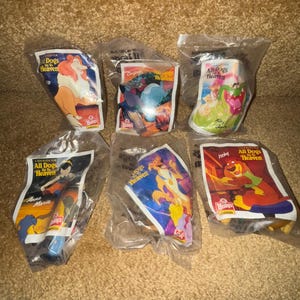 May include: Six plastic bags containing small plastic toys from the movie "All Dogs Go To Heaven". The bags are clear and show the toys inside. The toys are of different characters from the movie, including Charlie, Itchy, Anne Marie, and Carface. The bags are labeled with the movie title and the character's name.