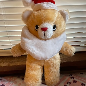 May include: A plush teddy bear wearing a red Santa hat with white trim and a white fur scarf. The bear is light brown and has black eyes and a red nose.