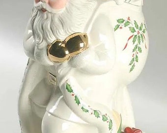 May include: A white ceramic Santa Claus figurine holding a bag of gifts. The bag is decorated with holly and red ribbon. The Santa is wearing a red hat with a white trim and a white beard.