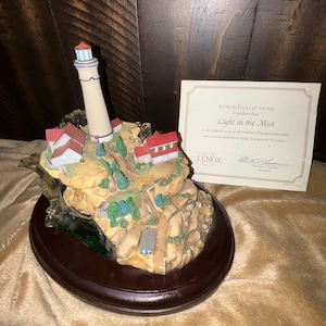 Può includere: Una statuetta in porcellana di un faro su una scogliera rocciosa con un piccolo villaggio. La statuetta è su una base in legno e viene fornita con un certificato di autenticità di Lenox Collections. Il certificato recita "Light in the Mist" ed è un'opera d'arte originale realizzata in porcellana legata e interamente dipinta a mano da Lenox.