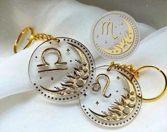 May include: Two circular clear acrylic keychains featuring gold-colored zodiac symbols for Libra and Leo. Each keychain displays a crescent moon, sun rays, and small stars. A separate circular graphic shows the Scorpio symbol with a moon and sun design.