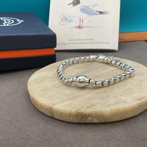 May include: A silver bracelet with a box chain design and a central spherical bead. The bracelet is displayed on a round, light-colored stone surface, with a navy blue box and a card featuring a bird illustration in the background.